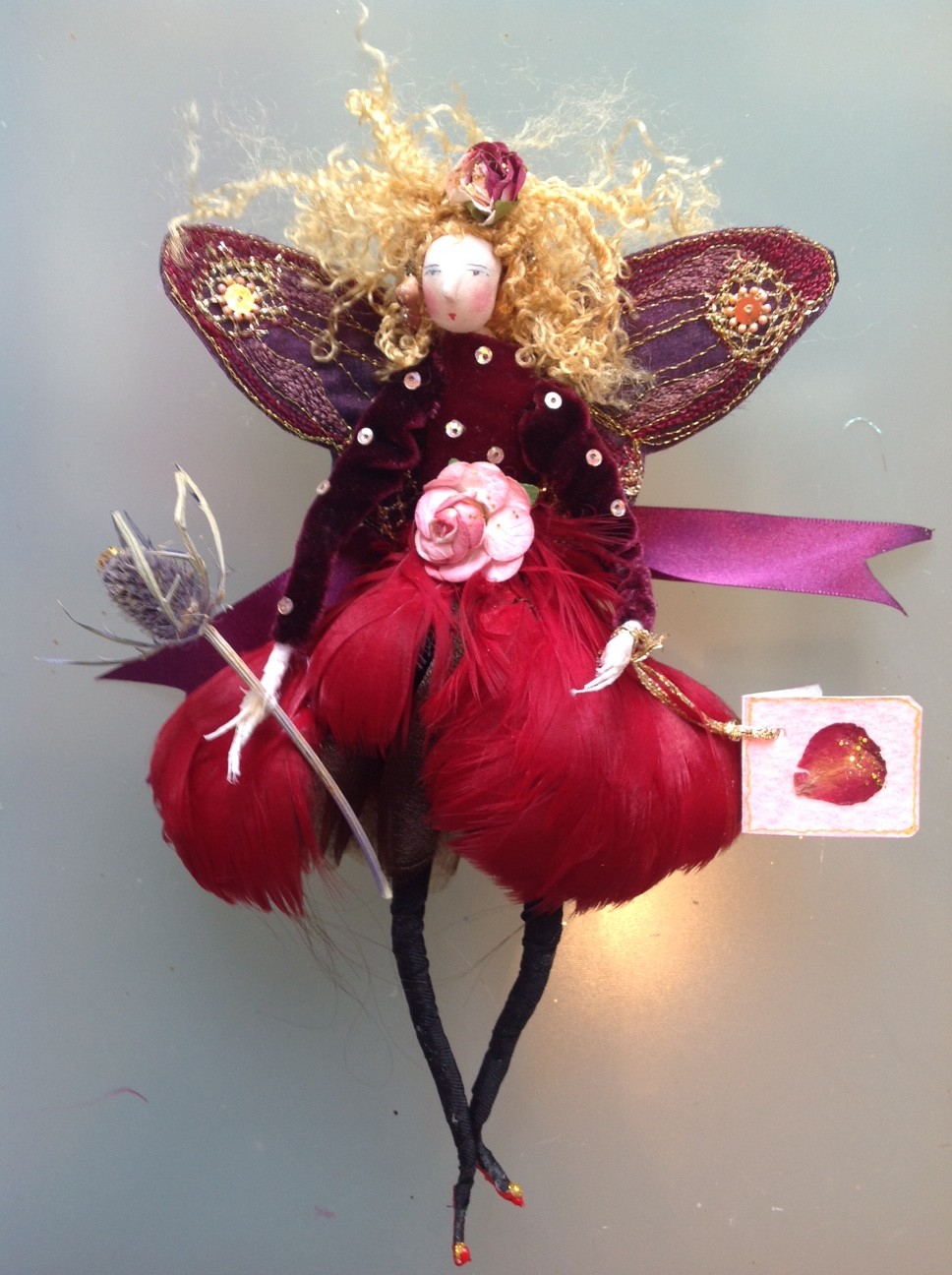 SARSAPARILLA, a 20cm fairy with a painted paper clay head. She is wearing a curled vintage feather and velvet dress with a paper rose at the waist. She has stiffly embroidered wings and is holding a dried sea holly. She comes in an organza bag with dried petals and her painted name tag. £40 plus £3 postage.
